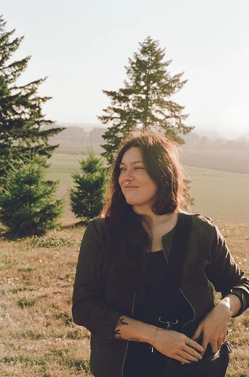 Jen Macias De La Parra standing outdoors in a field of young fir trees at golden hour, smiling and looking off to one side, with rolling farmland behind her.